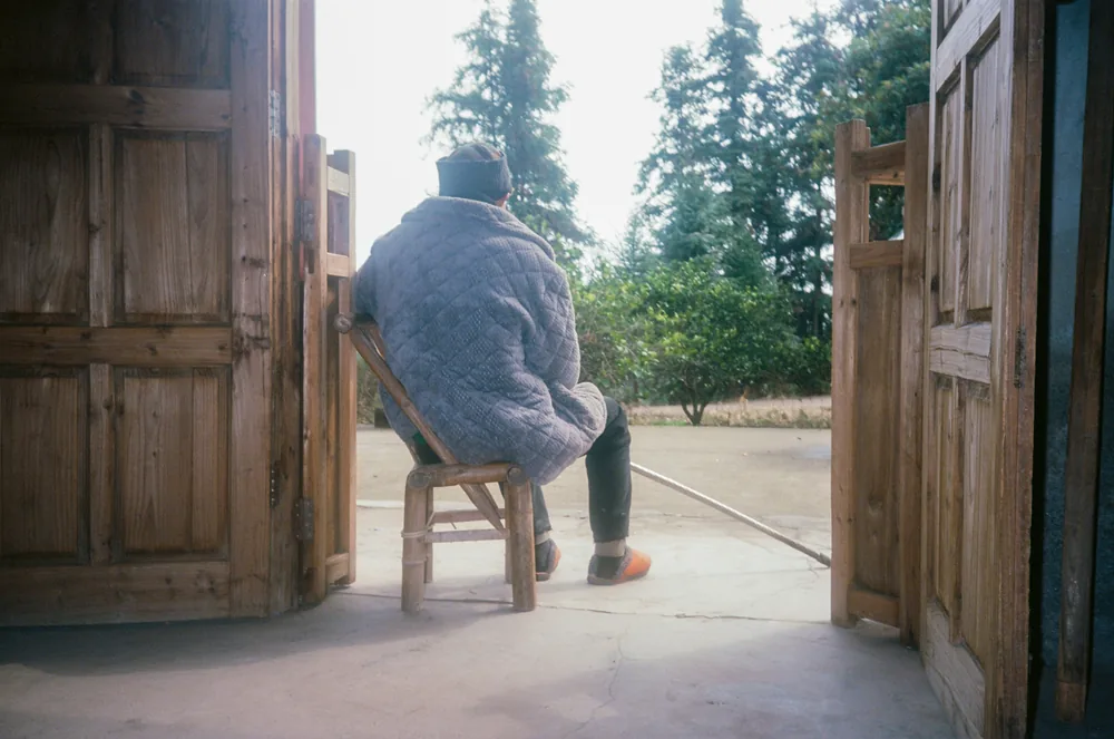 An older person sitting alone on a chair in a doorway, looking out at the yard.